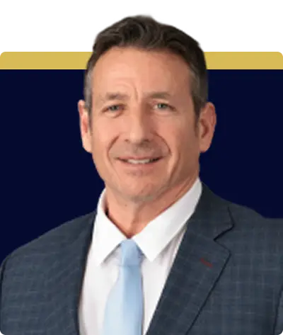 A middle-aged man wearing a checked dark suit, white shirt, and light blue tie stands against a dark blue background with a yellow stripe at the top. He has short brown hair and a slight smile.