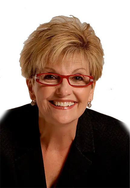 A smiling older woman with short, light blond hair, wearing red glasses and a black blazer, against a plain white background.